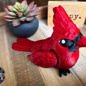 May include: A vibrant red cardinal figurine with black accents on the beak, eyes, and feet. The bird is perched on a wooden surface, with a succulent plant in a dark blue pot in the background. The figurine is a decorative item.