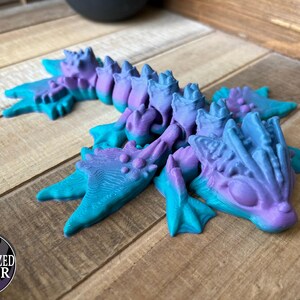 May include: A 3D-printed dragon figurine in shades of teal, purple, and blue. The dragon has a detailed, textured surface with a spiky spine and clawed feet. The figurine is resting on a wooden surface.