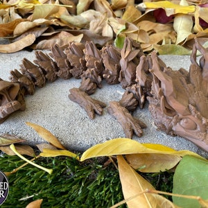 May include: A brown, 3D printed, dragon-like creature with a spiky, textured body and a long tail. The creature is lying on a gray surface with brown leaves scattered around it.