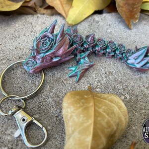 May include: A green and pink dragon keychain with a rose design. The dragon is attached to a silver key ring with a lobster clasp.