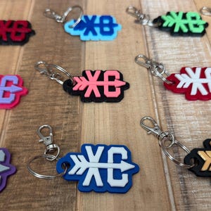 May include: A collection of multi-colored keychains featuring a layered design with a central arrow motif and the letters XC. Each keychain includes a metal split ring and a lobster clasp, showcasing various color combinations like blue, pink, green, and gold.