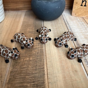 May include: Five miniature giraffe figurines with brown and white spotted patterns and black feet are arranged on a wooden surface. The giraffes are lying down, and a dark gray pot and a wooden sign are in the background.