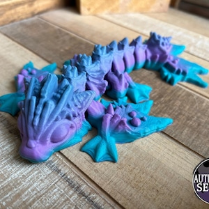 May include: A 3D-printed dragon figurine in shades of blue, purple, and teal. The dragon has a detailed, textured design with a spiky spine and fin-like appendages. The figurine is on a wooden surface, with an "AUTHORIZED SELLER" sticker.