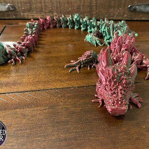 May include: A 3D printed dragon figurine with a segmented body. The dragon is painted in shades of red and green, with a metallic finish. The dragon's head is facing forward, with its mouth open and teeth visible.