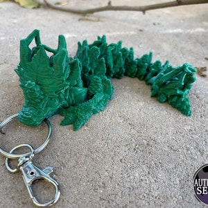May include: A green 3D printed dragon keychain with a silver key ring and clasp. The dragon is curled up with its head raised and its tail curled around its body.