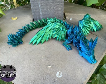 May include: A green and blue origami dragon with iridescent scales and wings. The dragon is lying on a gray concrete surface.