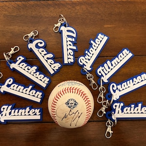 May include: Nine blue and white baseball-themed keychains with names printed on them, including Aiden, Brigdon, Caleb, Crayon, Hunter, Jackson, Kaiden, Landon, and Tillman. A baseball is in the center of the image.