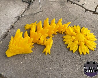 May include: A yellow 3D printed dragon figurine with a sunflower design. The dragon is lying on its side with its head resting on the ground. The sunflower design is on the dragon's back and tail.