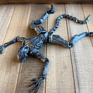 May include: A detailed, articulated, black and silver alien skeleton figure with a segmented tail. The figure is posed on a wooden surface.