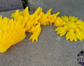 May include: A bright yellow 3D printed dragon figurine with a spiky back and a curled tail. The dragon is lying on a gray surface.