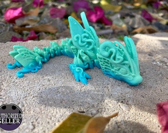 May include: A teal and blue 3D printed dragon figurine with intricate details. The dragon is lying on its side with its head resting on the ground. The figurine is made of a smooth, plastic-like material.