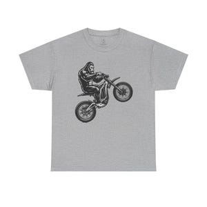 Puede incluir: Una camiseta gris jaspeado de manga corta con un estampado gráfico en blanco y negro de un gorila montando una moto de cross mientras hace un caballito. El diseño está centrado en el pecho de la prenda, mostrando al primate en una pose dinámica.