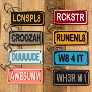 May include: Eight colorful keychains with different words and phrases printed on them. The keychains are made of plastic and have a metal ring for attaching to keys. The words on the keychains are: LCNS PL8, RCKSTR, CROOZAH, RUNENL8, DUUUUUDE, W8 4 IT, AWESUMM, WH3R MI.