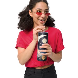 May include: A cylindrical tumbler featuring a dark blue night sky design with a large white crescent moon, a full moon, and small stars. The lower portion of the tumbler displays a colorful floral arrangement with orange, pink, and blue flowers.