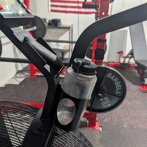 May include: Black and red exercise bike with a clear water bottle attached. The water bottle is black and clear. The bike is in a gym setting with weights and an American flag in the background.