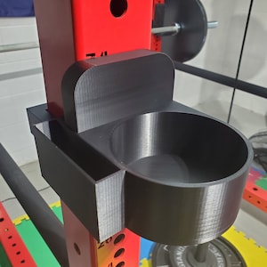 May include: A black 3D printed holder with a cup and a small compartment attached to a red metal weightlifting rack.
