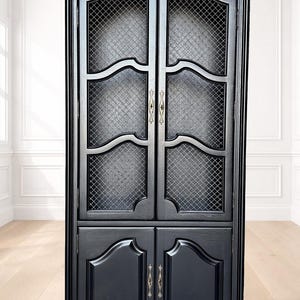 May include: A tall black cabinet with an arched top, featuring two upper doors with a diamond-patterned metal mesh grid and two solid lower doors. The cabinet includes ornate brass-toned hardware on each door and a smooth, dark finish.
