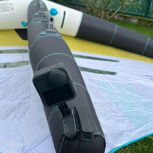Peut inclure: Une voile de planche à voile noire et verte avec une caméra noire et grise montée sur le wishbone. La caméra est orientée vers l'avant et est fixée au wishbone à l'aide d'une sangle noire.