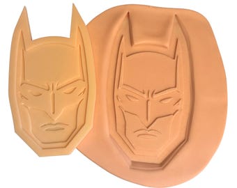 May include: A light peach-colored silicone mold featuring a recessed Batman mask design alongside a matching light peach-colored Batman mask cutout. The mold has a smooth, flexible surface with a clear, debossed outline of the character's face.