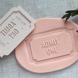 May include: Two white silicone moulds shaped like movie ticket stubs. The top mould reads "ADMIT ONE" and the bottom mould reads "TIMOA ONE".