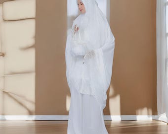 May include: A full-length image of a person wearing a white prayer garment. The garment includes a head covering, a long skirt, and a matching handbag. The outfit is adorned with lace detailing, creating a modest and elegant appearance.