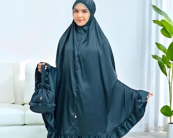 May include: A dark teal prayer garment with a matching drawstring bag. The garment features a head covering with lace trim and a ruffled hem. The bag has a gold-colored emblem. The text "Rattanmerch" is visible at the bottom right.