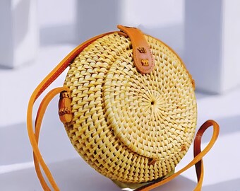 May include: A round, woven rattan handbag with a light brown leather strap and accents. The bag is a natural beige color and features a circular design. The strap is a warm orange-brown color. The bag is a stylish accessory.