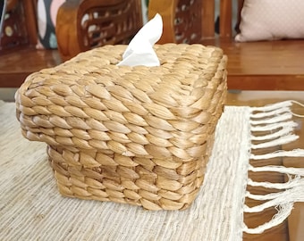 May include: A woven wicker tissue box holder with a white tissue peeking out of the top. The holder is sitting on a light brown woven mat with white fringe.