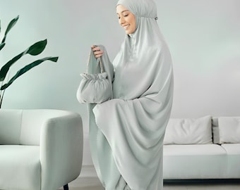 May include: A light green prayer garment set, including a head covering and matching dress, is held with a small purse. The outfit is made of a soft, flowing fabric. The text "Rattanmerch" is visible at the bottom right.