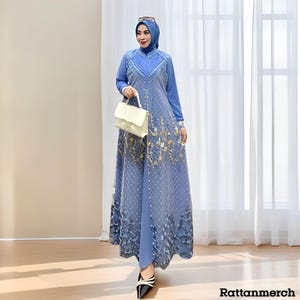 Op de afbeelding: Een lange, vloeiende blauwe jurk met gouden bloemaccenten en bijpassende hoofdbedekking. De jurk heeft lange mouwen en een gedetailleerd lijfje. Een crèmekleurige handtas wordt in de hand gehouden. De tekst "Rattanmerch" is rechtsonder zichtbaar.