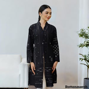 Op de afbeelding: Een zwarte jas met lange mouwen en een wit geometrisch patroon. De jas heeft een V-hals en een knoopsluiting. De tekst "Rattanmerch" staat rechtsonder.