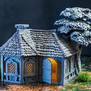 Könnte beinhalten: Ein Miniatur-Grau-Cottage mit einem strukturierten Dach, einem Schornstein und einem Baum daneben. Das Cottage hat eine blaue Tür und Fensterrahmen. Das Innere leuchtet mit warmem Licht, was auf eine gemütliche Atmosphäre hindeutet. Das Gesamtthema ist Fantasy.