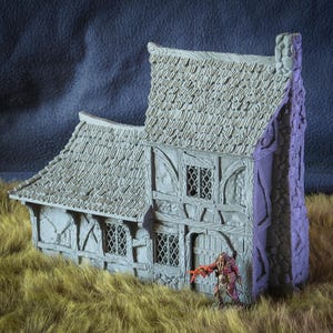 Könnte beinhalten: Ein graues, miniatures, zweistöckiges, strohgedecktes Haus mit einem Steinkamin und einer Holztür. Das Haus hat ein kleines Fenster im zweiten Stock und zwei Fenster im ersten Stock. Eine Miniaturfigur mit einem roten Umhang und einem Schwert steht vor dem Haus.