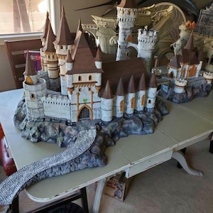 May include: A detailed model of a white and brown castle with a stone bridge leading to the front entrance. The castle has multiple towers and a stone wall surrounding the main structure. The model is set on a rocky base with green and brown accents.