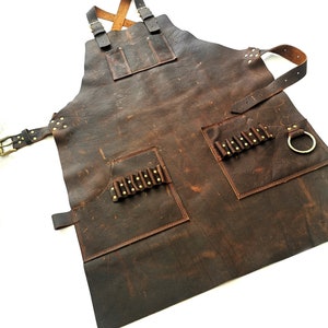 May include: A brown leather apron with multiple pockets and a brass ring. The apron has a distressed look and is secured with straps.