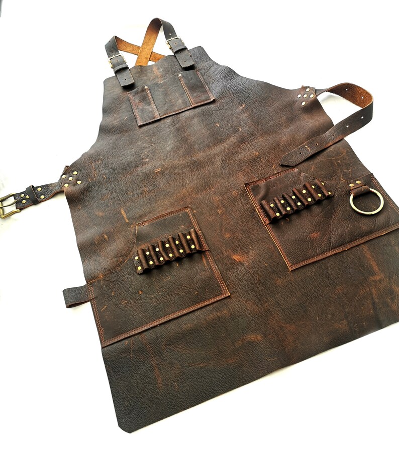 May include: A brown leather apron with multiple pockets and a brass ring. The apron has a distressed look and is secured with straps.