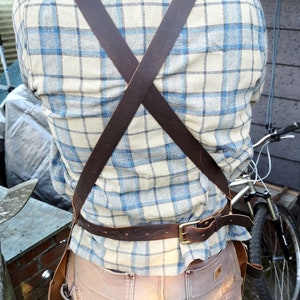 May include: A brown leather apron with adjustable straps and a belt. The apron is worn over a plaid shirt and brown pants.