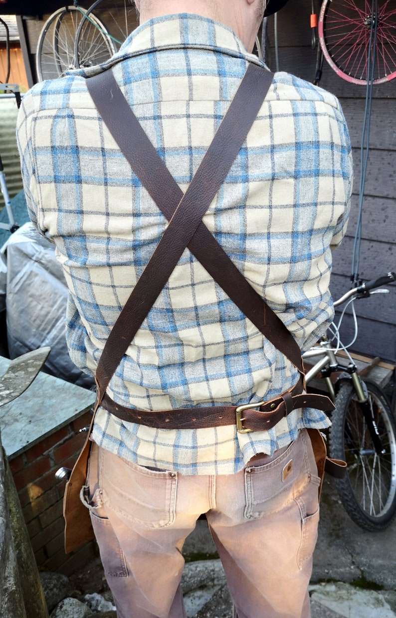 May include: A brown leather apron with adjustable straps and a belt. The apron is worn over a plaid shirt and brown pants.