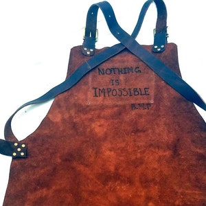 May include: Brown leather apron with adjustable straps and a brass buckle. The apron has the text "NOTHING IS IMPOSSIBLE" written on it.