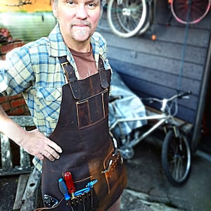 May include: A man wearing a plaid shirt and a brown leather apron with multiple tool pockets. He is standing in front of a wooden wall with a bicycle in the background. The apron has a variety of tools in the pockets, including screwdrivers and wrenches.