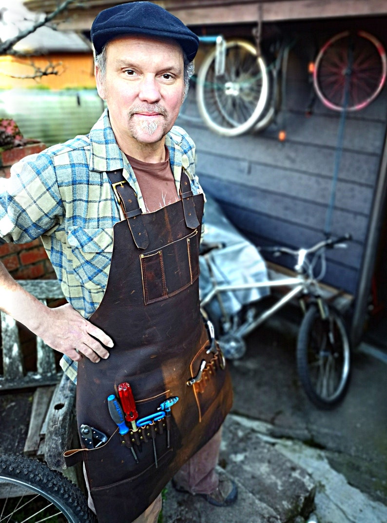 May include: A man wearing a plaid shirt and a brown leather apron with multiple tool pockets. He is standing in front of a wooden wall with a bicycle in the background. The apron has a variety of tools in the pockets, including screwdrivers and wrenches.