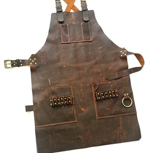 May include: A brown leather apron with multiple pockets and a brass buckle. The apron has a criss-cross strap design and a ring on the side.