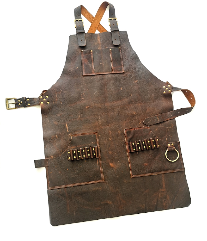 May include: A brown leather apron with multiple pockets and a brass buckle. The apron has a criss-cross strap design and a ring on the side.