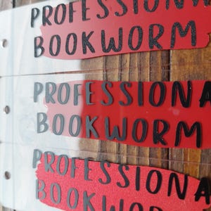 Könnte beinhalten: Drei transparente Acryl-Lesezeichen mit den Worten "PROFESSIONAL BOOKWORM" in schwarzer Schrift auf rotem Hintergrund. Die Lesezeichen werden auf einer Holzoberfläche präsentiert.