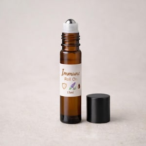 Könnte beinhalten: Braune Glas-Roll-on-Flasche mit silberner Kugel und schwarzem Deckel. Das Etikett trägt die Aufschrift "Immune Roll On" mit einem Schild, Lavendel und einer Tropfenillustration. Die Flasche enthält 15 ml Produkt.