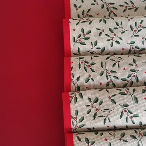 Puede incluir: Camino de mesa navideño plegado con un estampado de acebo. El corredor tiene un fondo beige con hojas verdes y bayas rojas. Los bordes están ribeteados con un borde rojo liso.