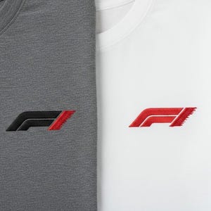 Op de afbeelding: Twee T-shirts, één grijs en één wit, elk met een geborduurd Formule 1-logo. Het logo is zwart en rood op het grijze shirt en rood op het witte shirt. De shirts zijn gemaakt van een zachte, getextureerde stof. Het logo is ongeveer 5 cm breed.