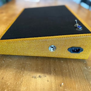 May include: A yellow pedalboard with a black top and a power cord and a 1/4 inch jack on the side.