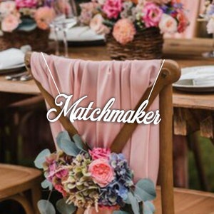 May include: A pink fabric chair sash with the word "Matchmaker" written in white cursive letters. The chair is decorated with a bouquet of pink, purple, and green flowers.