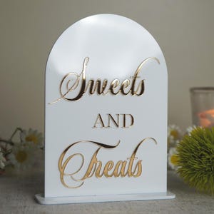 Może przedstawiać: Biały, łukowaty znak ze złotym napisem "Sweets AND Treats". Znak znajduje się na stole z niebieskim kieliszkiem do wina, kwiatami i zapaloną świecą w tle. Dekoracyjny przedmiot na imprezę.